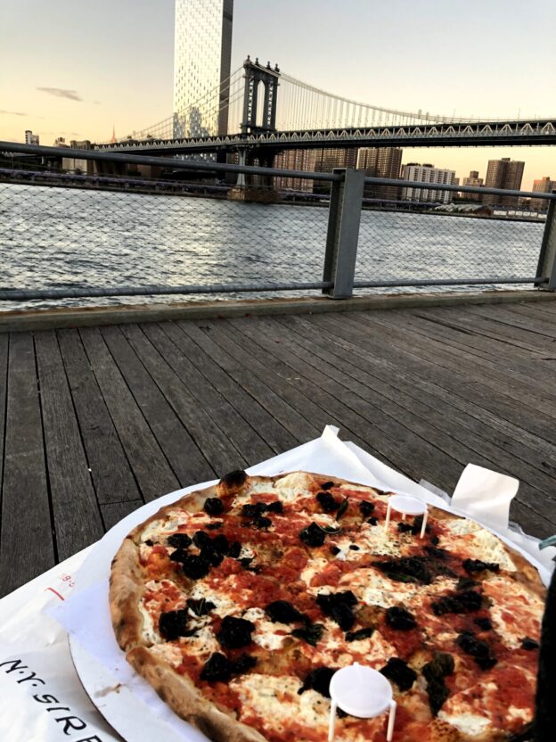 image of a large pizza margherita with olives. In the background, the Brooklyn Bridge at sunset.